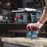 A carpenter sanding a wood piece with a sander in his woodshop full of power tools
