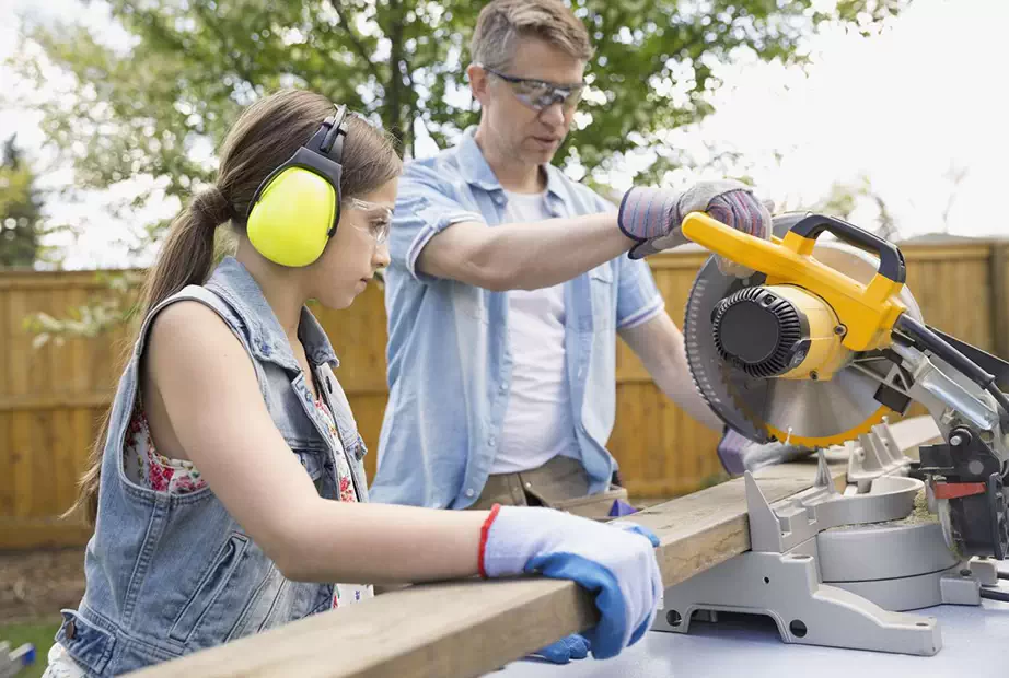 A man and a girl are cutting a wood piece with miter saw