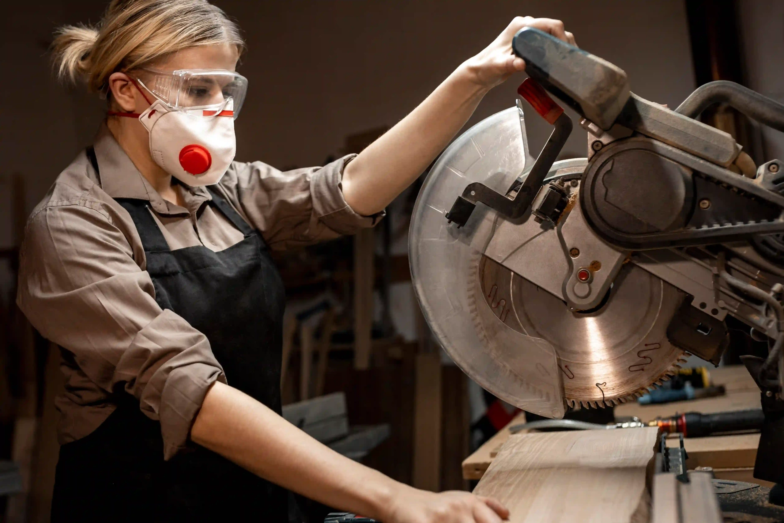 A side view of a female carpenter wearing safety gear, operating a circular saw in a woodworking workshop.