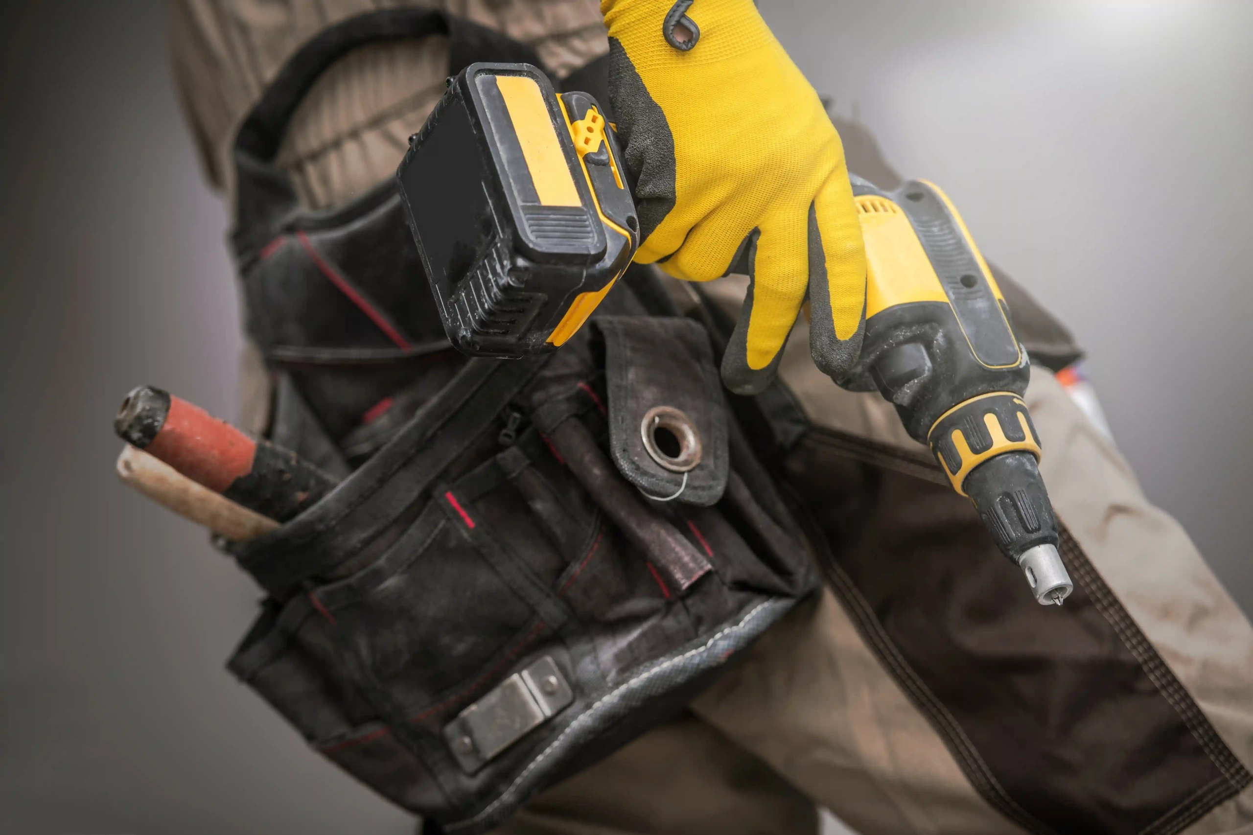 A close-up of a constructor wearing a tool belt, holding a cordless drill in his gloved hand.