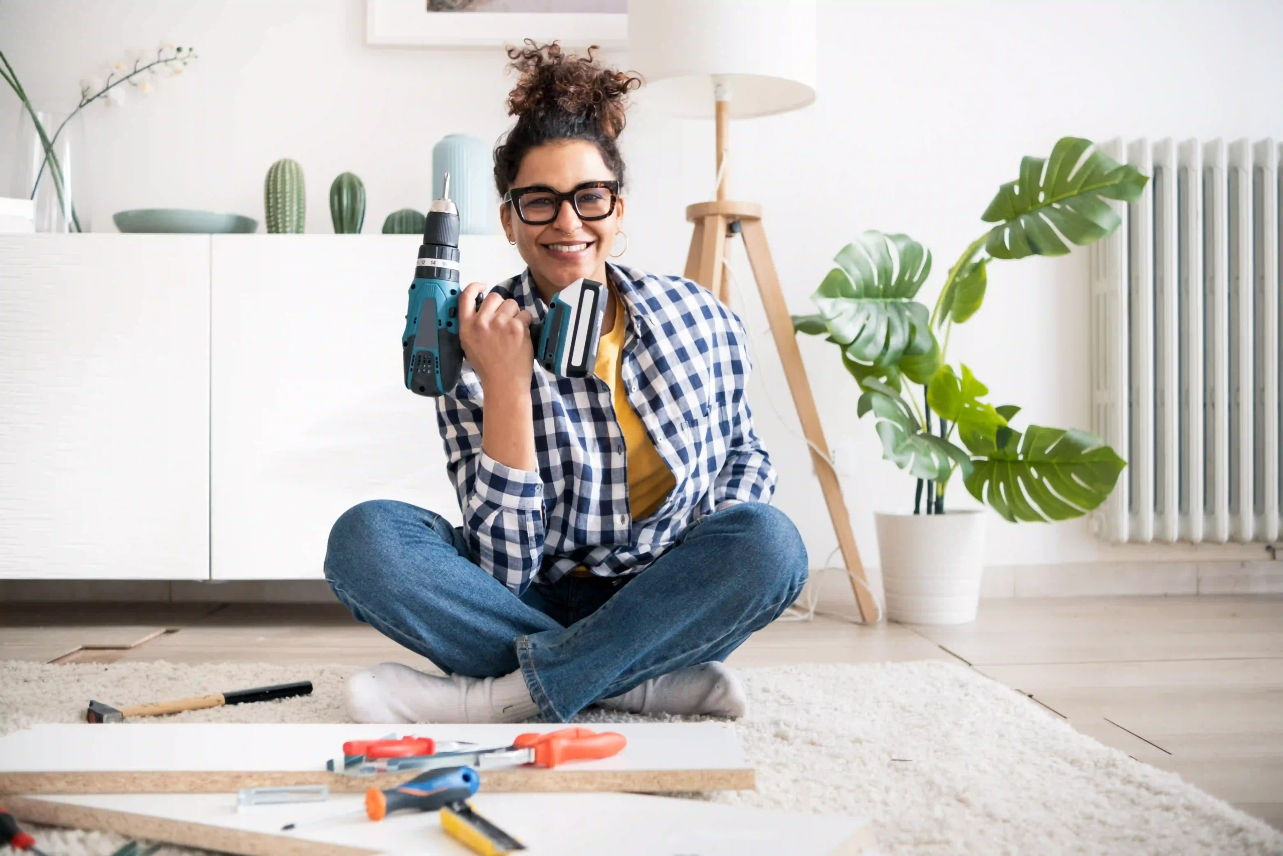 A smiling woman is holding a cordless drill, surrounded by tools for a DIY project.