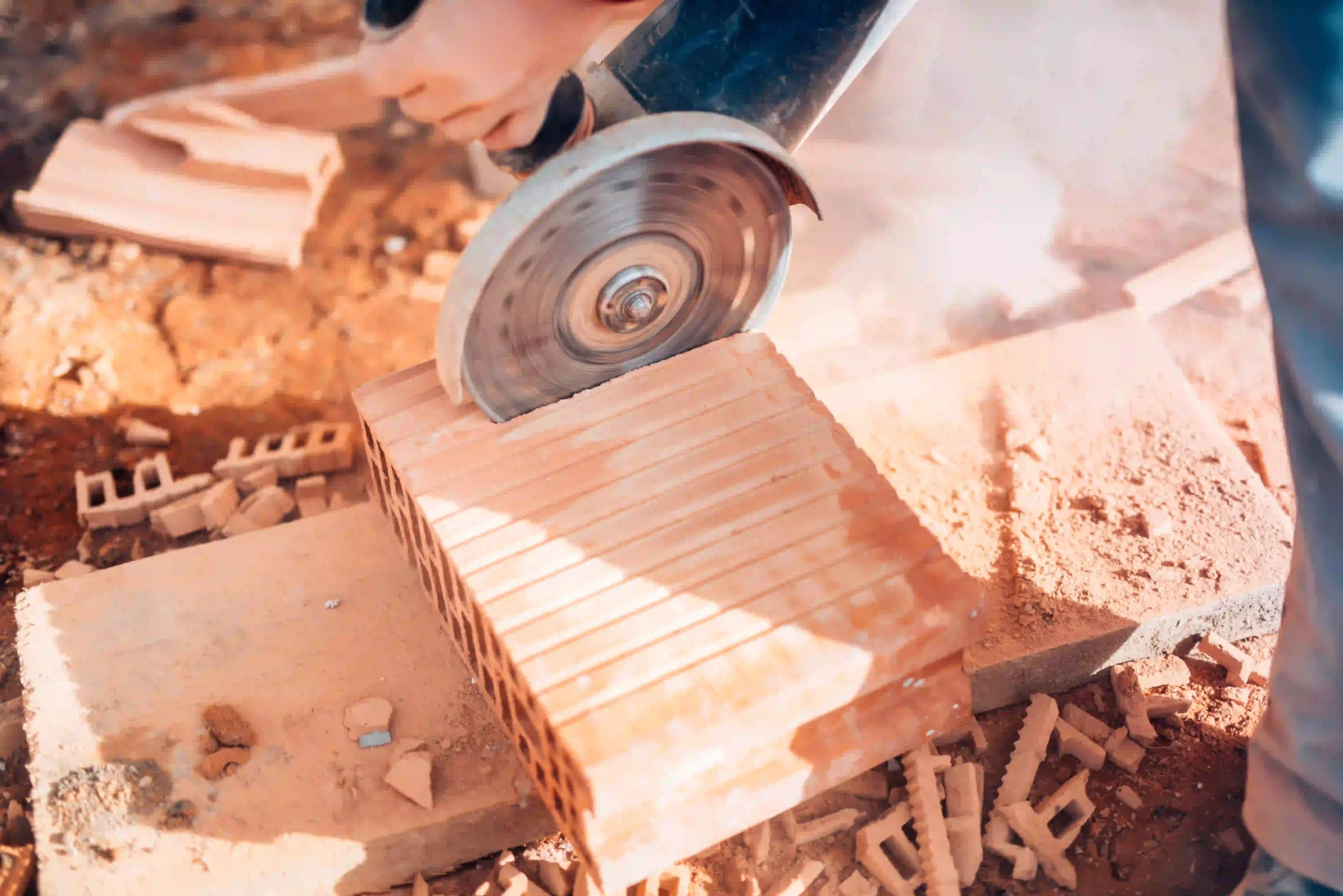 A worker using a cordless angle grinder to cut a brick in an outdoor construction setting.