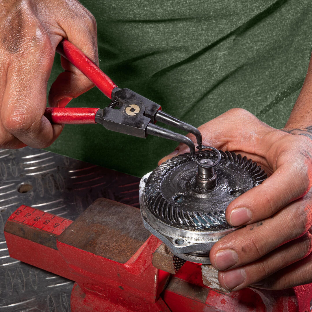 Mechanic using a Ronix bent snap ring pliers with red handles to remove a circlip from a greased gear assembly secured in a red bench vise.
