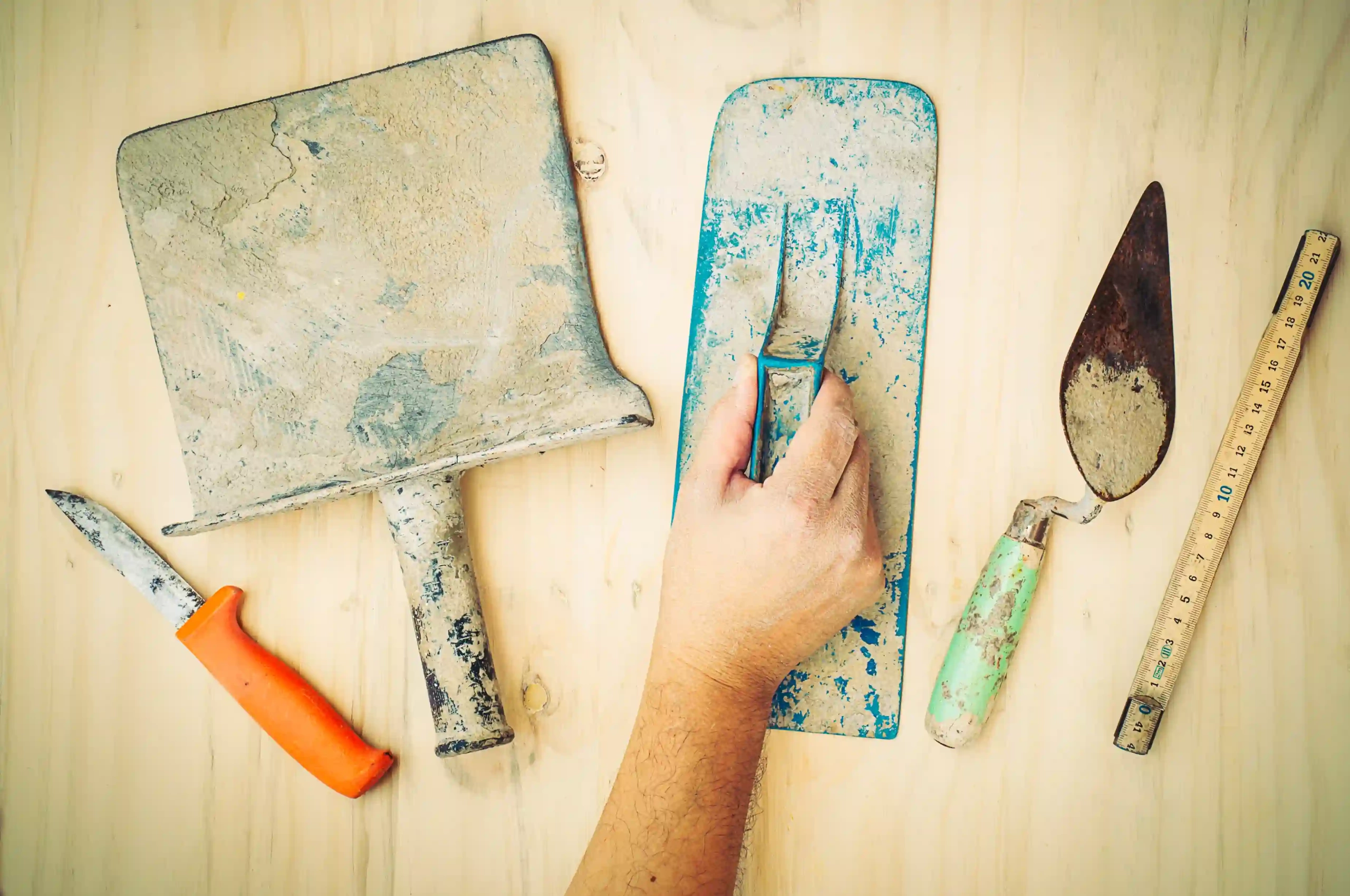 Dirty masonry tools on a wooden surface, including trowels, a knife, and a ruler. A worker's hand grips a blue float.