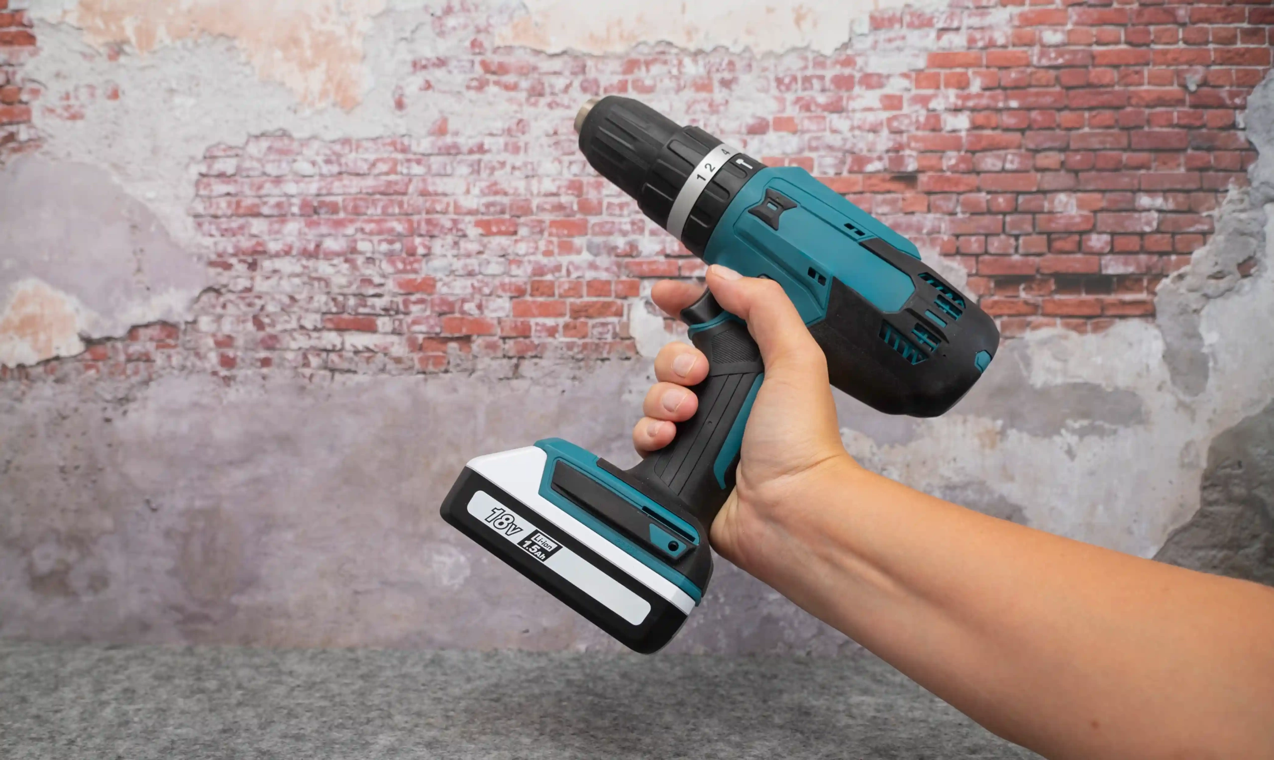 Hand holding 18V cordless drill against brick wall background